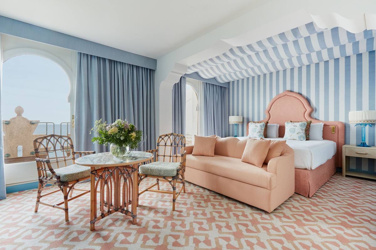 A hotel room with a large window, a round table with chairs, a sofa, and a bed with a unique headboard.