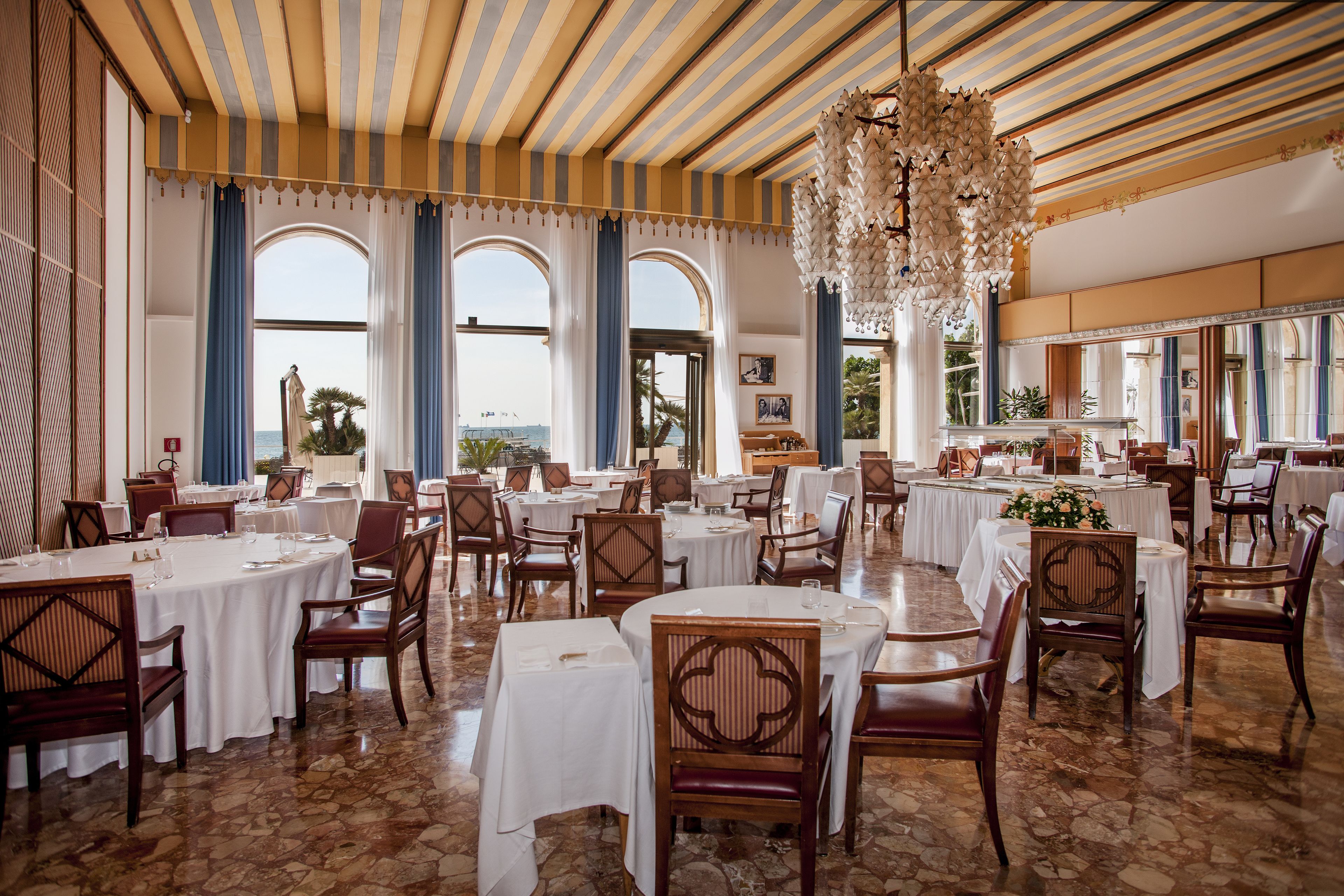 Un vaste espace dédié à la salle à manger, avec des tables et des chaises rondes, de grandes fenêtres ornées de rideaux bleus et une vue sur la mer.