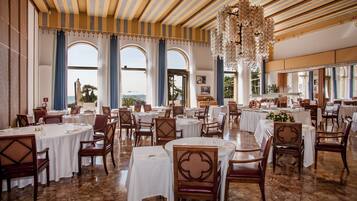 Un vaste espace dédié à la salle à manger, avec des tables et des chaises rondes, de grandes fenêtres ornées de rideaux bleus et une vue sur la mer.