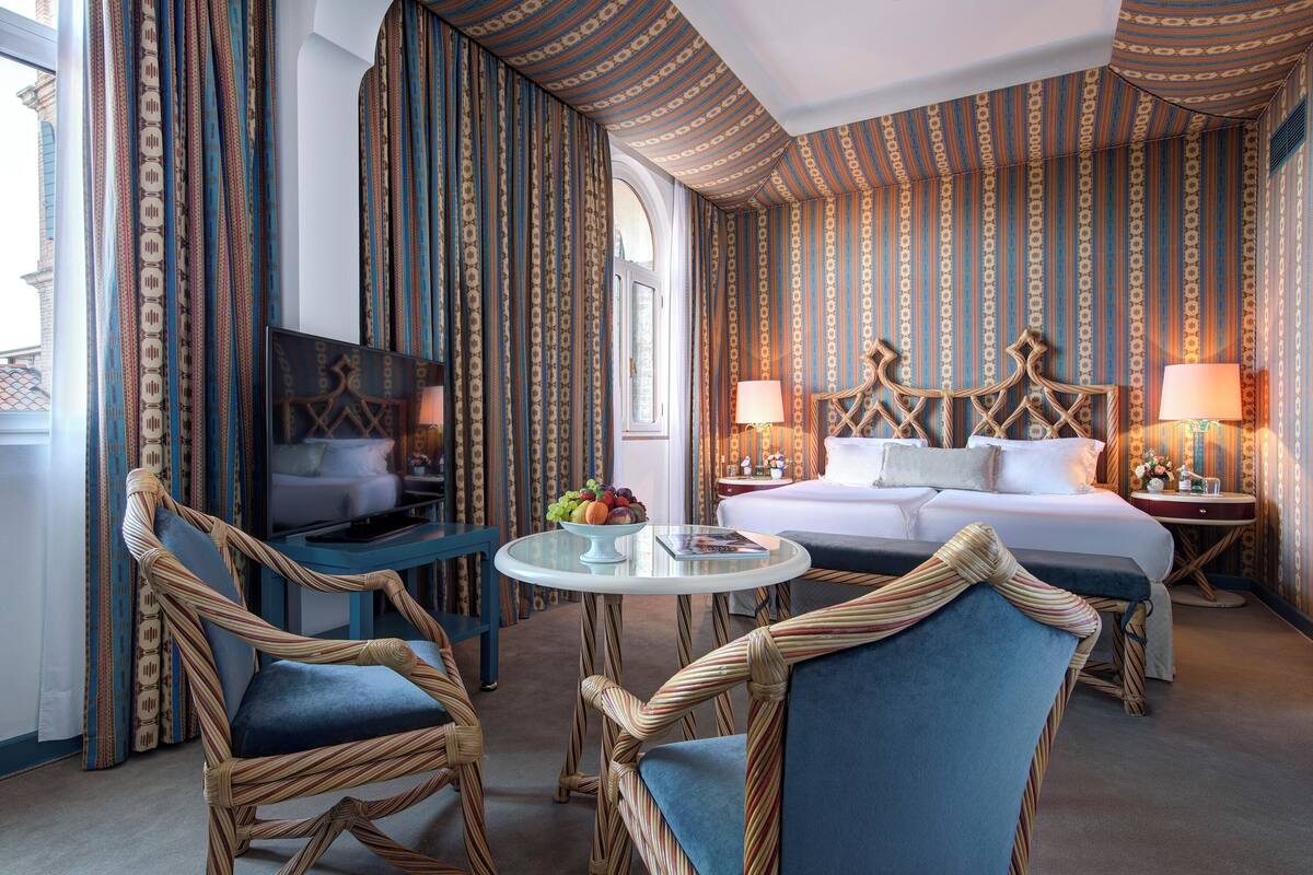 A hotel room with a bed, two nightstands, a TV, a small table with fruit, and wicker chairs.