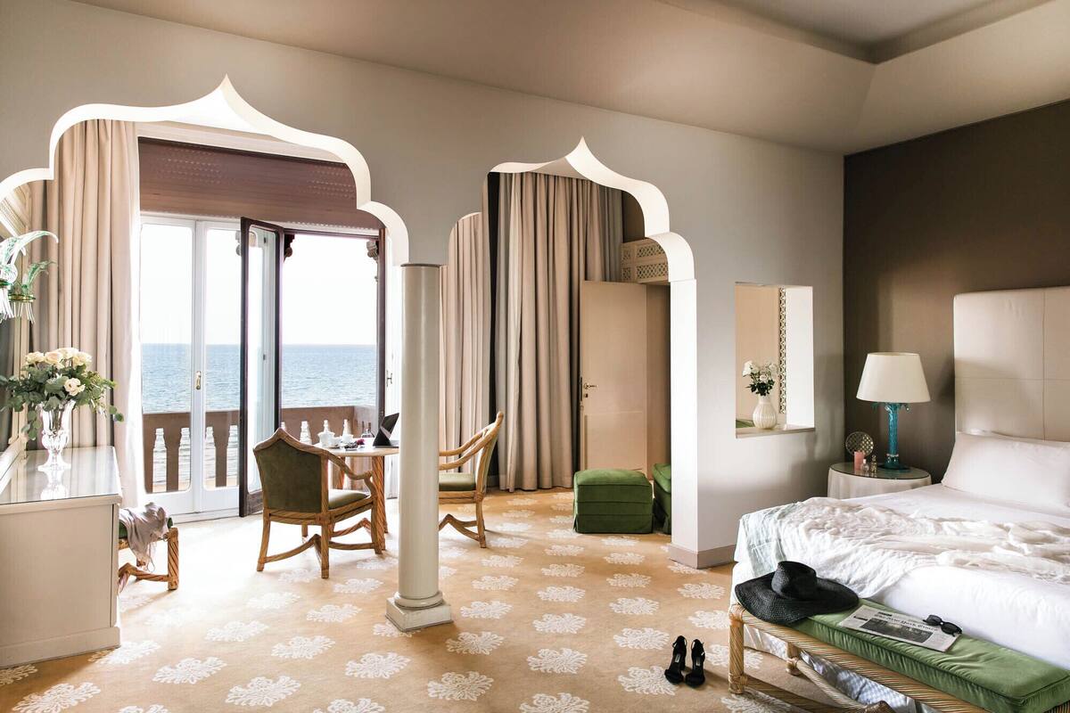 A hotel room with a bed, a desk with a chair, a small table, and a view of the sea through an open door.