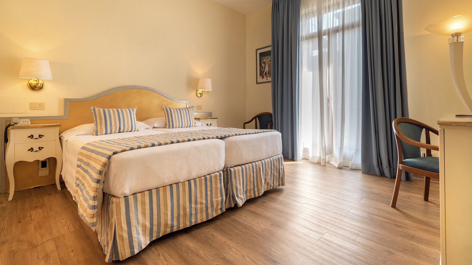 Cama doble con ropa de cama a rayas, mesitas de noche con lámparas, un escritorio con silla y un ventanal con cortinas.