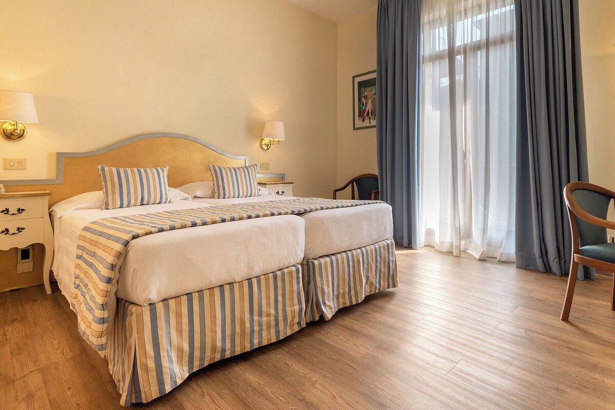 A double bed with striped bedding, bedside tables with lamps, a desk with a chair, and a large window with curtains.