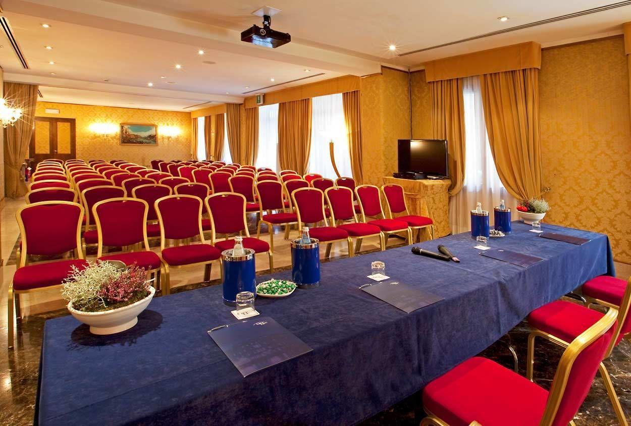 Un salón de conferencias con hileras de sillas rojas, una mesa larga con mantel azul, un televisor y un proyector en el techo.
