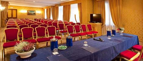 Un salón de conferencias con hileras de sillas rojas, una mesa larga con mantel azul, un televisor y un proyector en el techo.