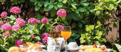 A table set with breakfast items including eggs, toast, and pastries, with two glasses of tea and juice, surrounded by lush greenery and pink flowers.