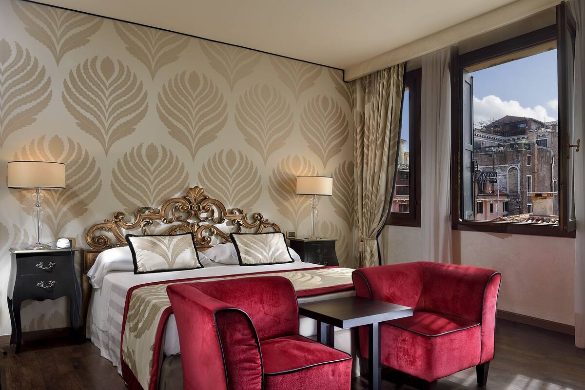 Une chambre d’hôtel avec un grand lit, deux tables de chevet avec des lampes, un fauteuil rouge rembourré, une petite table et une fenêtre donnant sur la ville.