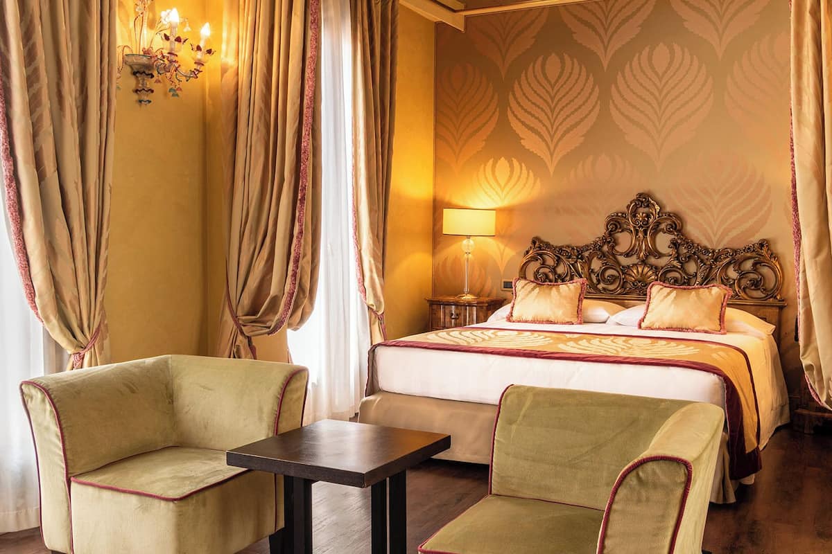 Une chambre d’hôtel avec un grand lit, deux fauteuils, une petite table et une table de chevet avec une lampe. Les murs sont recouverts d’un papier peint à motifs et la pièce est dotée de rideaux.