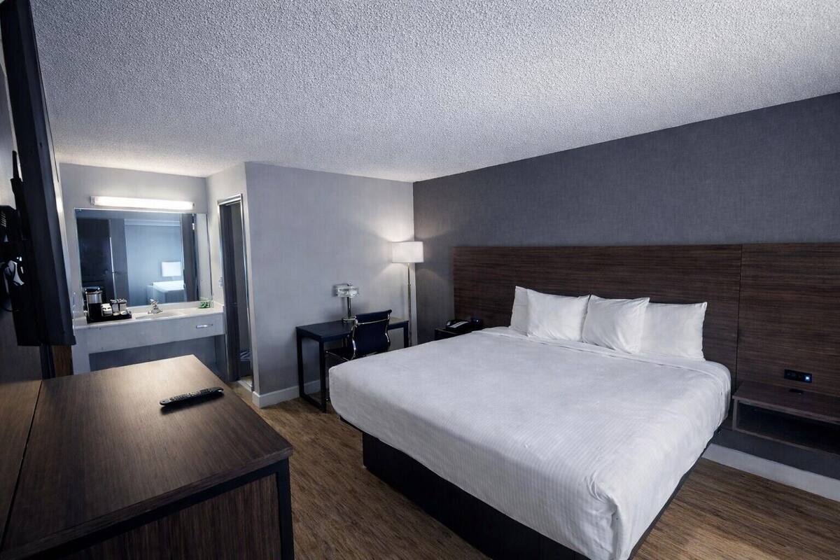 A modern hotel room with a large bed, a desk, a chair, a nightstand, a lamp, and a mirror.