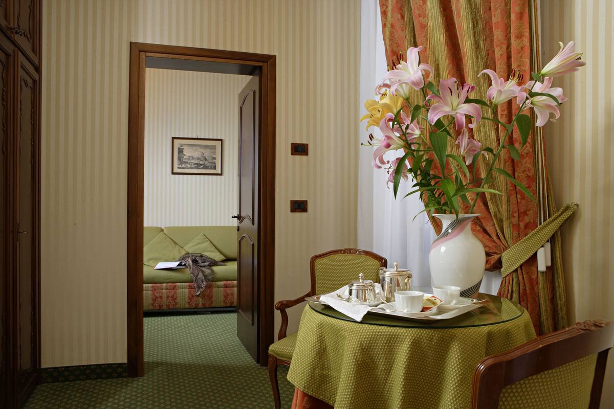 A room with a dining table set for tea, a green patterned sofa, and a vase with flowers.