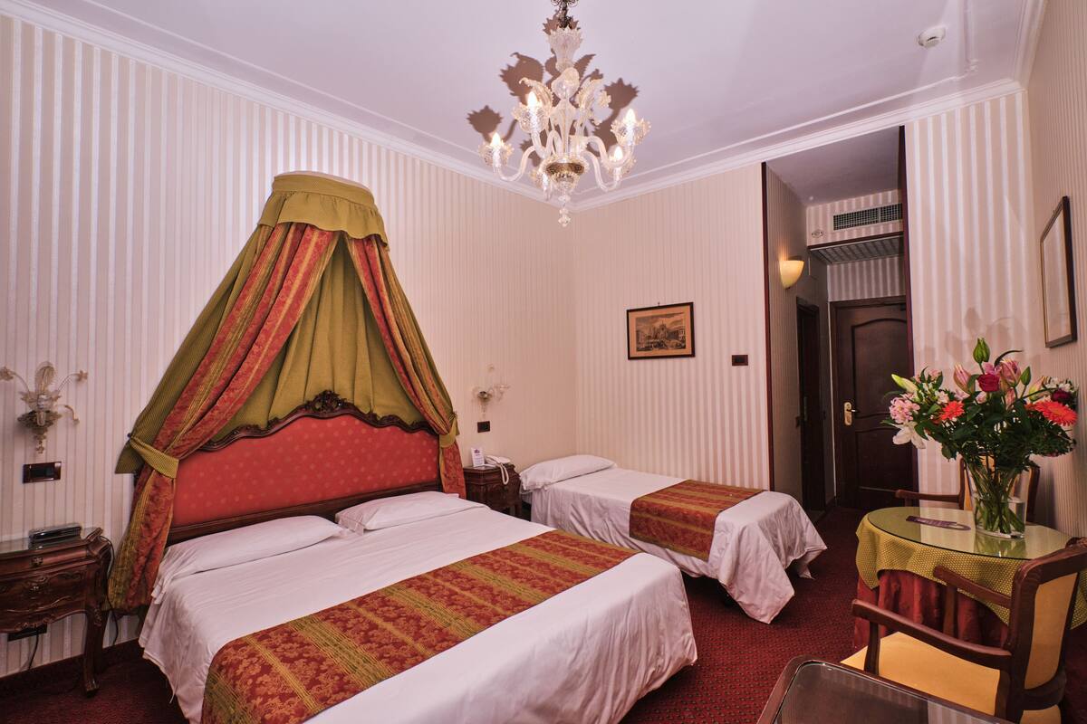 A hotel room with two beds, a chandelier, a small table with a vase of flowers, and a door leading to another room.