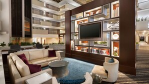 A modern hotel lobby with a high ceiling, multiple floors, a large flat-screen TV, a sofa, a coffee table, and a bookshelf.