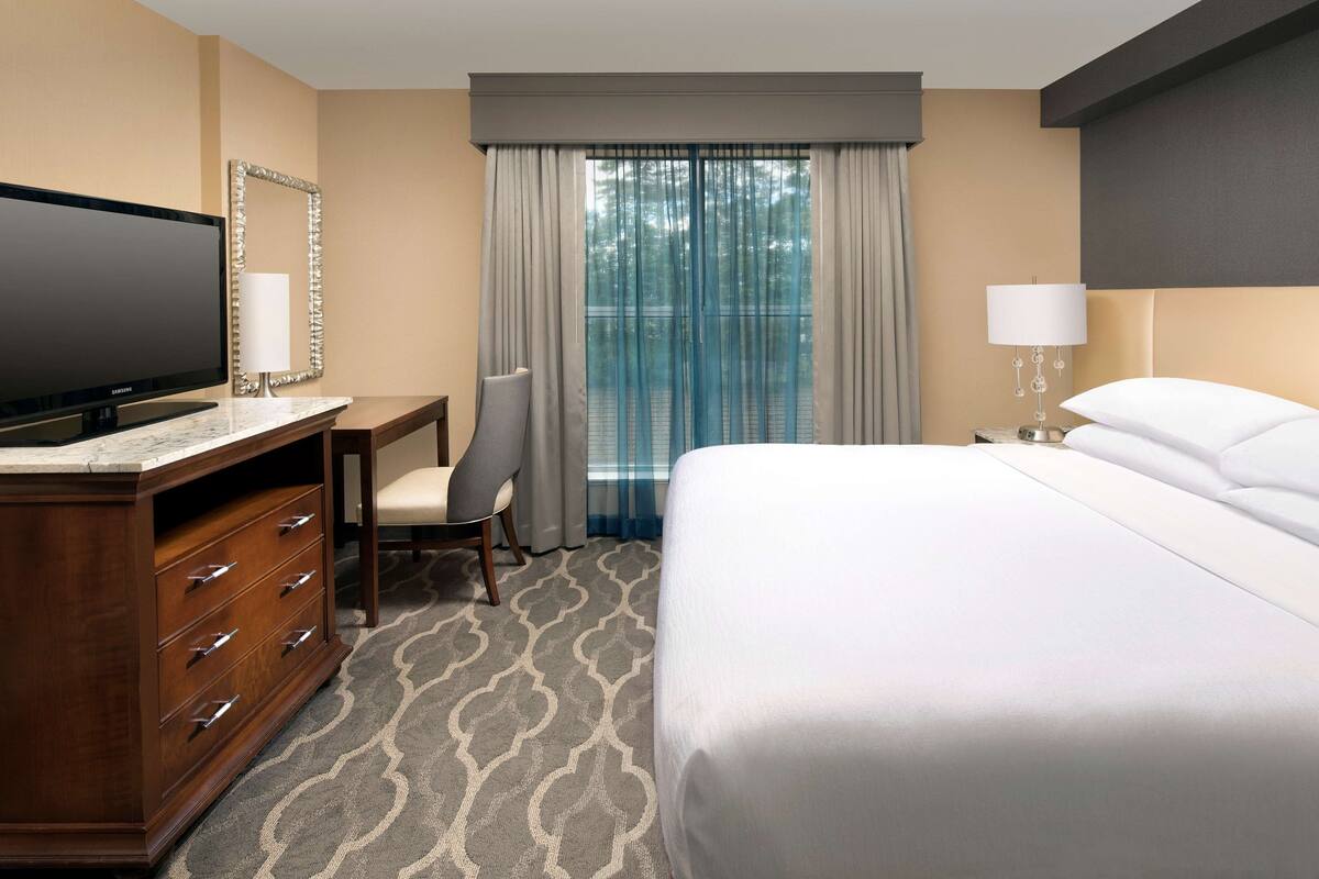 A hotel room with a bed, a desk with a TV, a chair, a lamp, and a window with curtains.