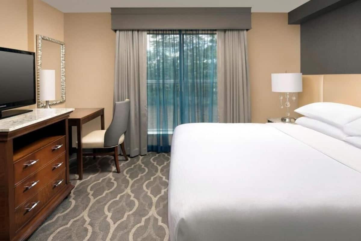 A hotel room with a bed, a desk with a television, a chair, a dresser, and a window with curtains.