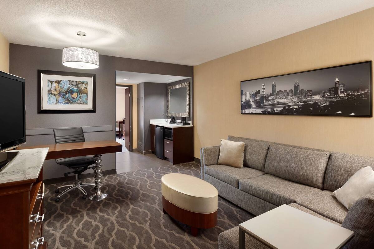 A hotel room with a dining area, a sofa, a coffee table, a TV, a desk with a chair, a large framed cityscape picture, and a patterned carpet.