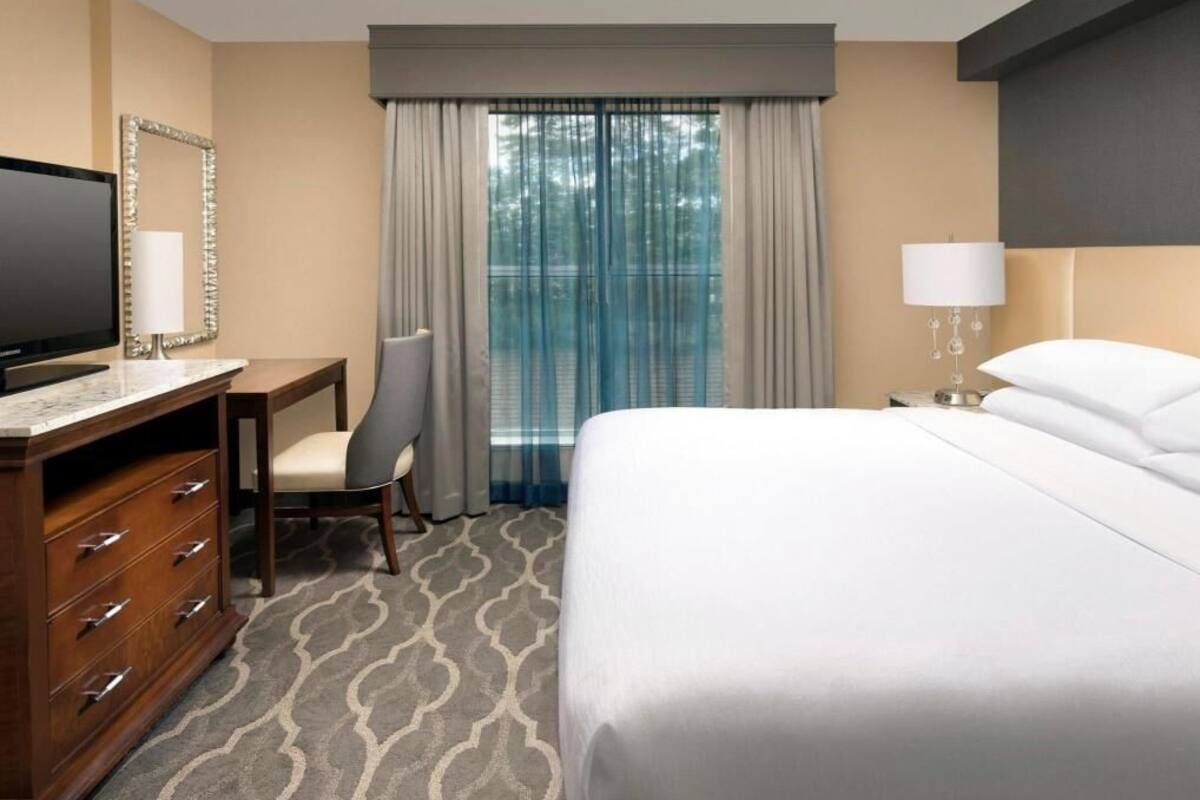 A hotel room with a bed, a desk with a television, a chair, a dresser, and a window with curtains.