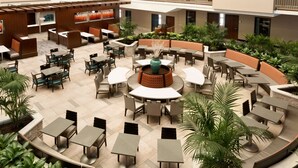 A spacious outdoor patio area with multiple seating arrangements, including round tables and booths, surrounded by lush greenery and potted plants.