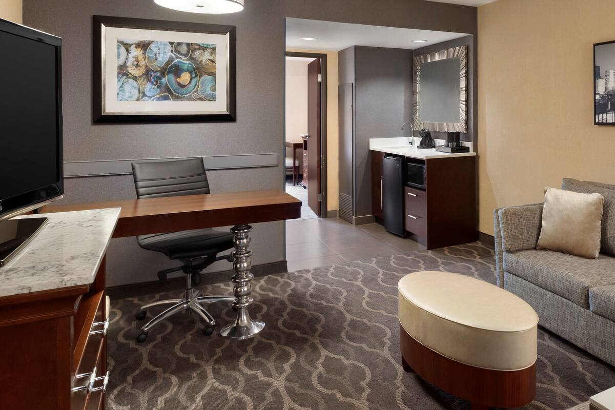 A modern hotel room with a dining area, a sofa, a television, and a bathroom with a mirror.