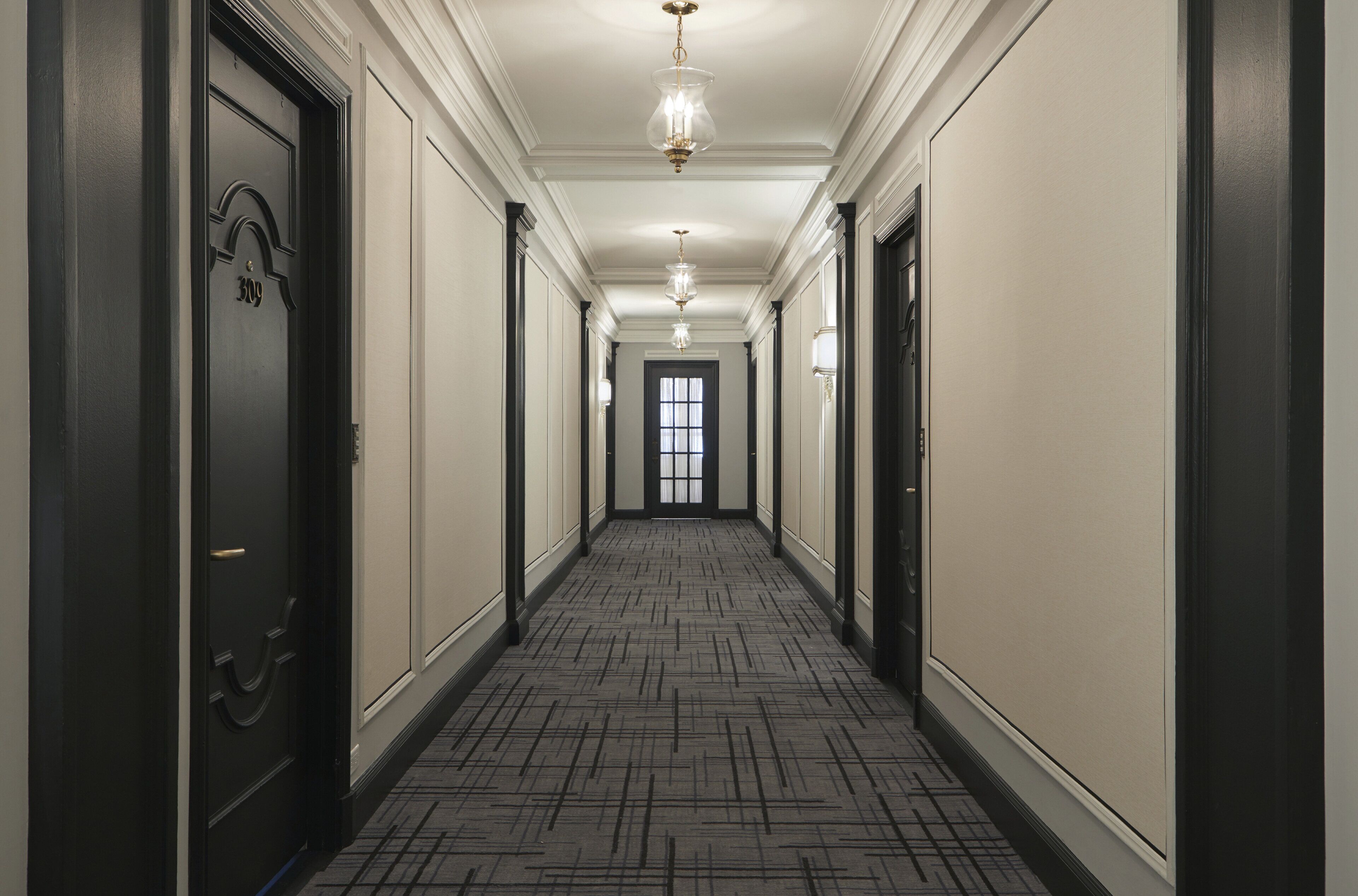 A long, narrow hallway with a patterned carpet, black doors, and a chandelier.