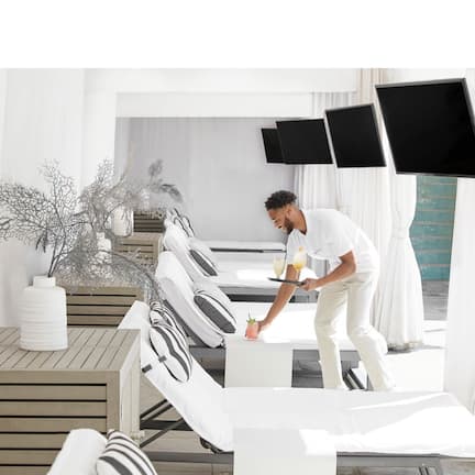 A modern hotel room with a wooden floor, white furniture, a potted plant, and a flat-screen TV on the wall.