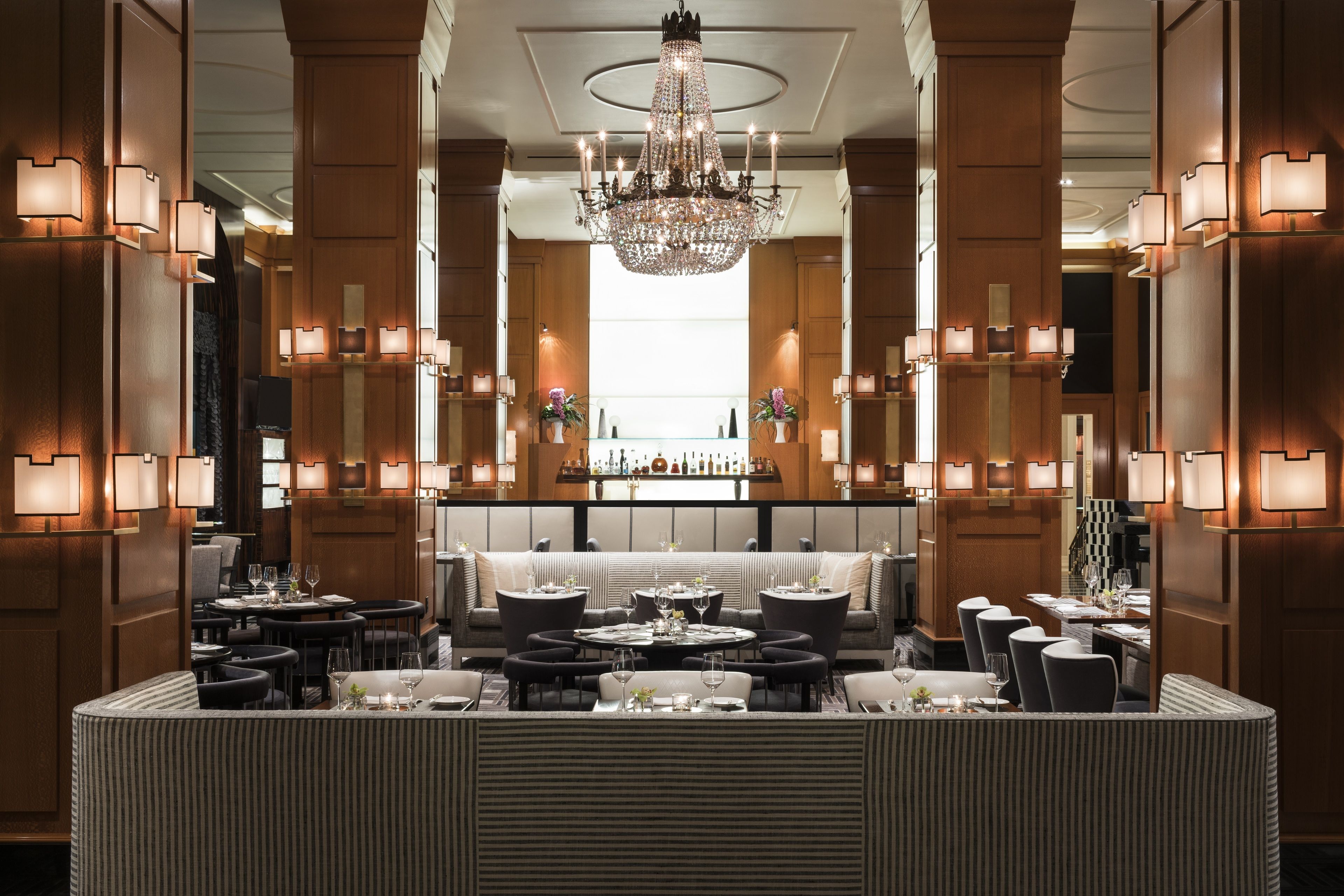 A luxurious hotel lobby with a central bar, elegant chandeliers, and a seating area with round tables and chairs.