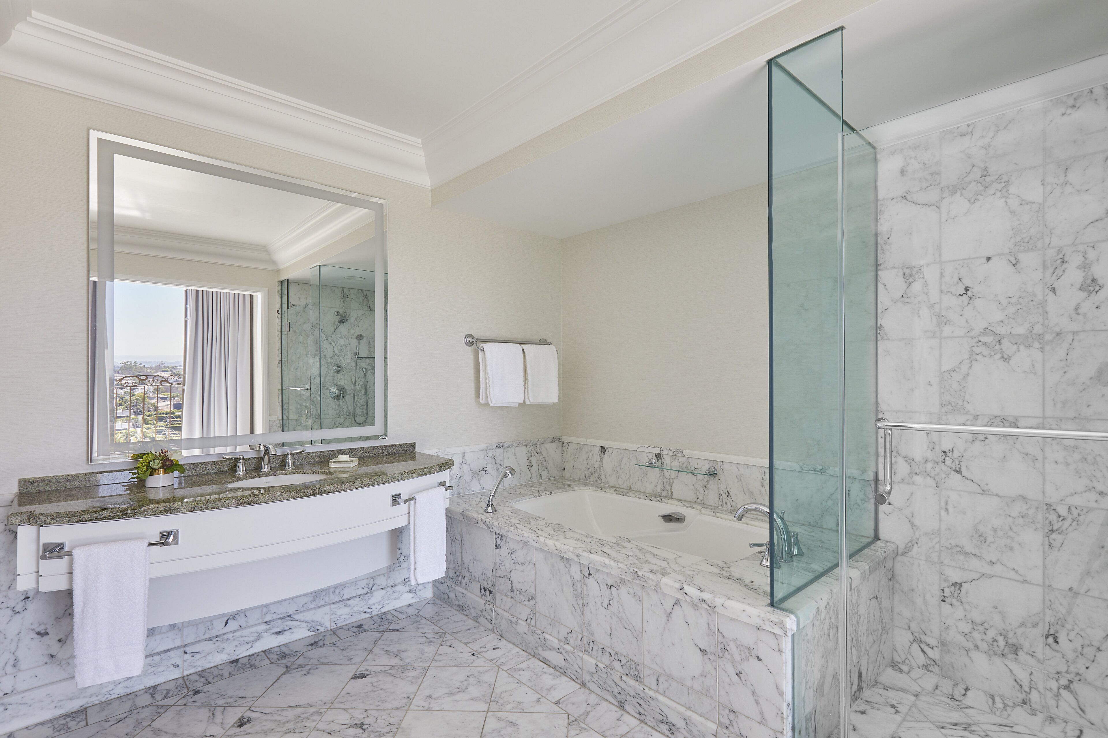 A modern bathroom with a marble sink, a large mirror, a glass-enclosed shower, and a view of the city.