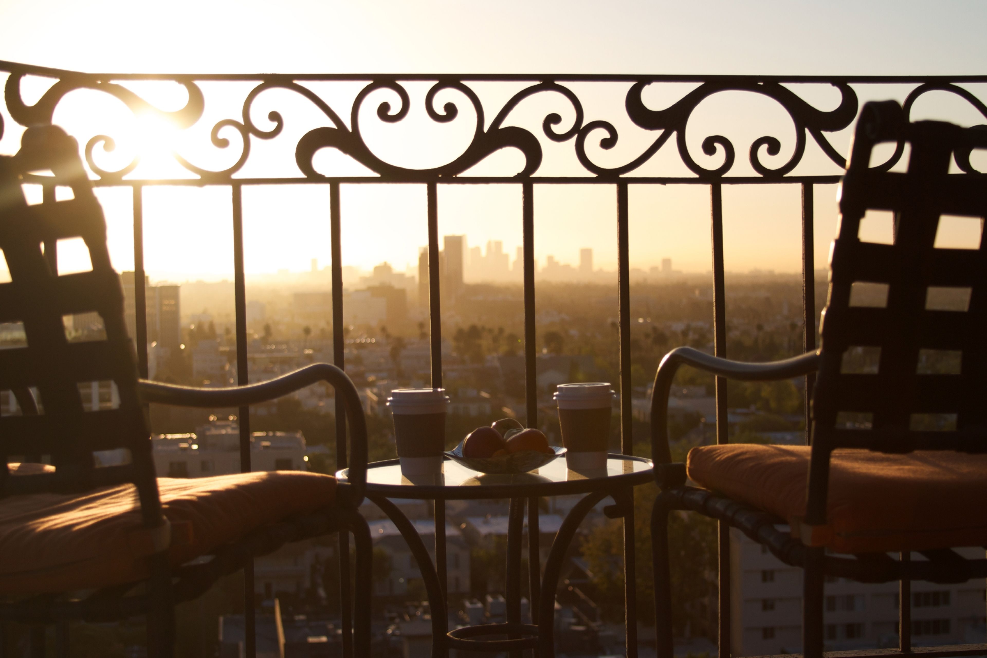 Un balcón con vistas a un paisaje urbano al atardecer. Hay dos sillas, una mesa, dos tazas y un plato de comida.