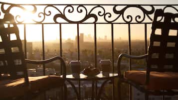 Un balcón con vistas a un paisaje urbano al atardecer. Hay dos sillas, una mesa, dos tazas y un plato de comida.