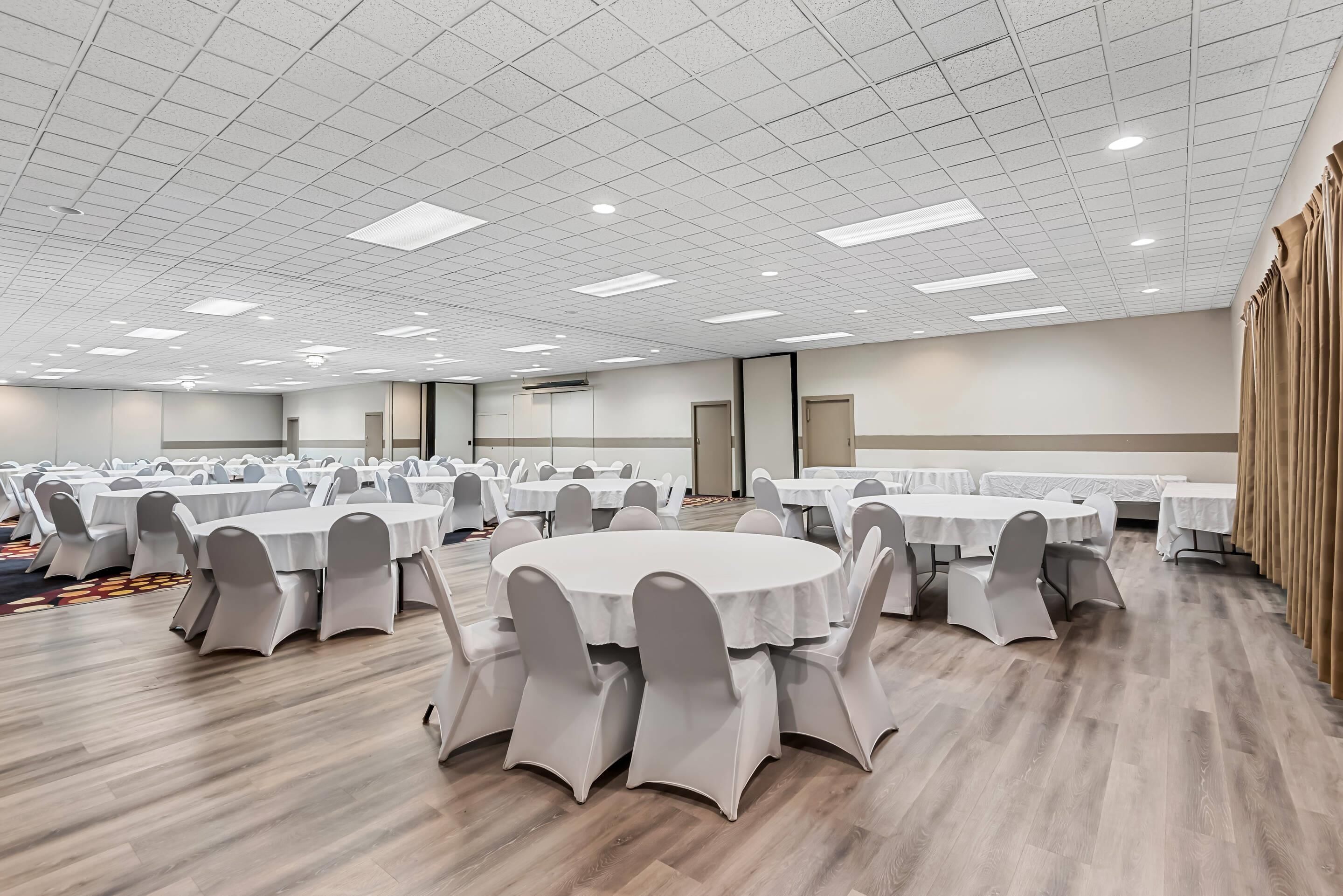 A large conference room with multiple round tables and chairs, equipped with ceiling lights and a wooden floor.