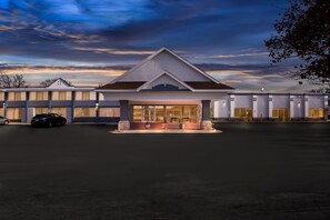 A hotel with a central entrance, a parking lot, and a row of buildings in the background.