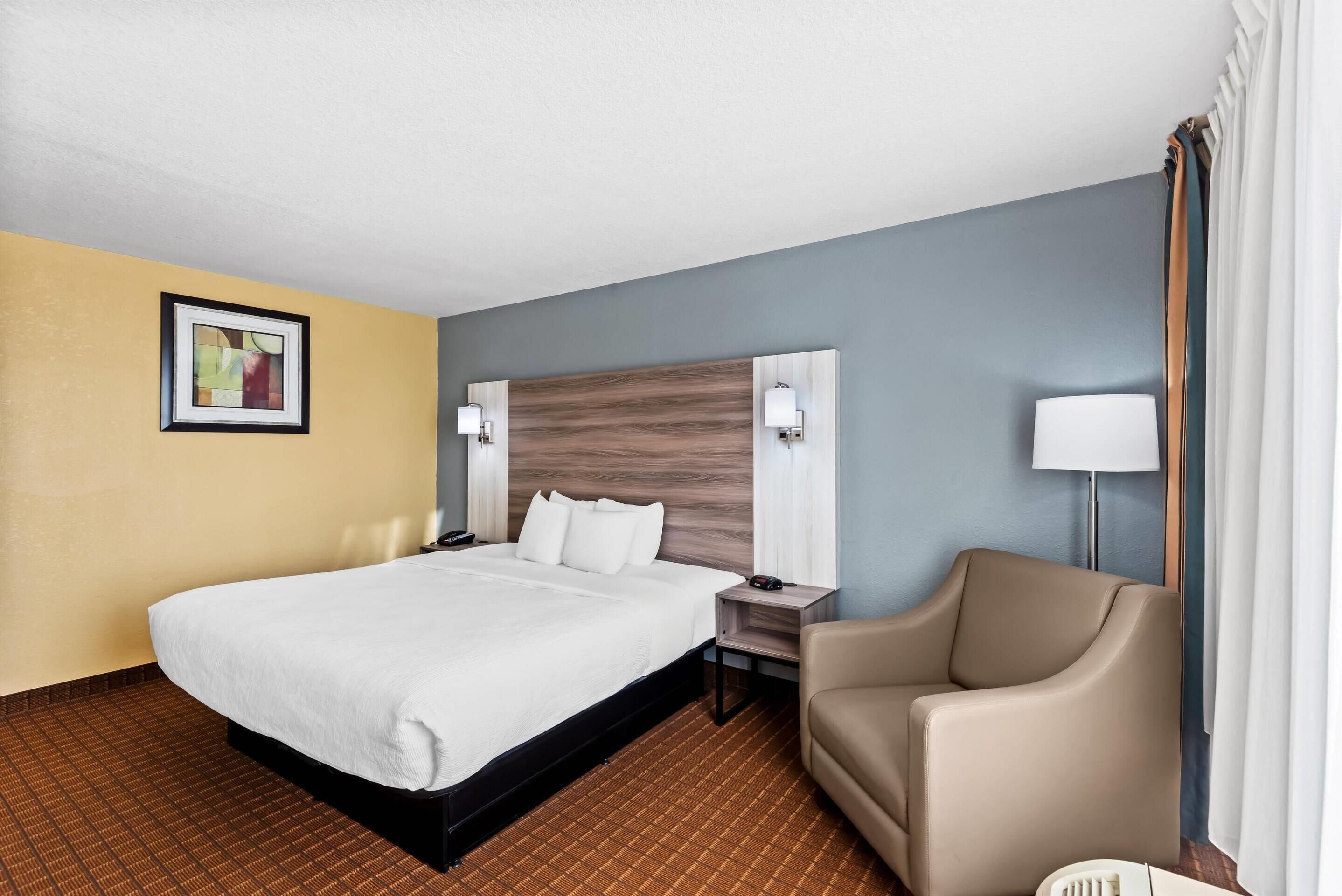 A hotel room with a large bed, a chair, a nightstand, a lamp, and a painting on the wall.