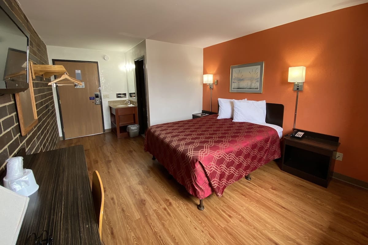 A hotel room with a wooden floor, a bed with a red patterned bedspread, a nightstand with a lamp, a wooden dresser, and a bathroom visible through an open door.