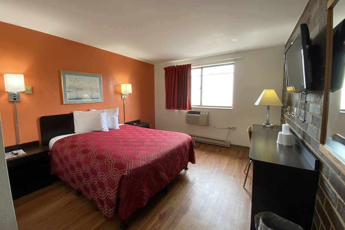 A hotel room with a large bed, two bedside lamps, a flat-screen TV mounted on the wall, a window with red curtains, and an air conditioning unit.