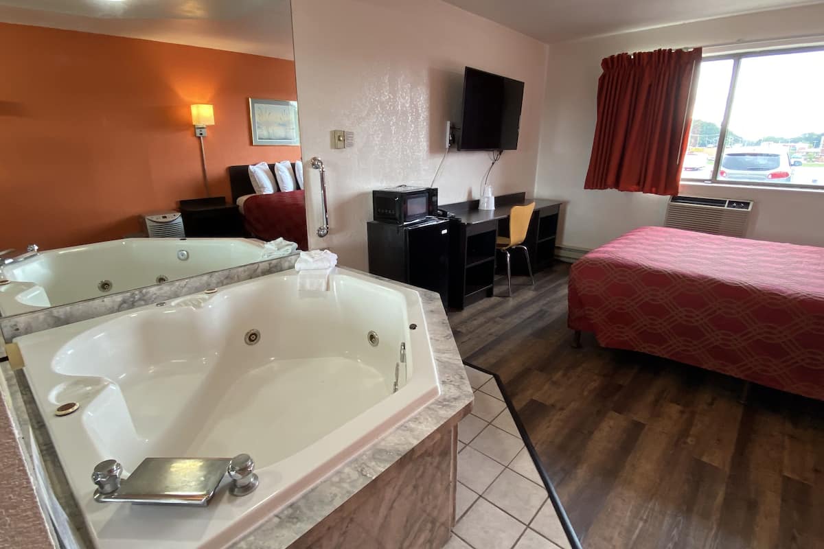 A hotel room with a large bathtub, a desk with a chair, a bed, a TV mounted on the wall, and a window with curtains.