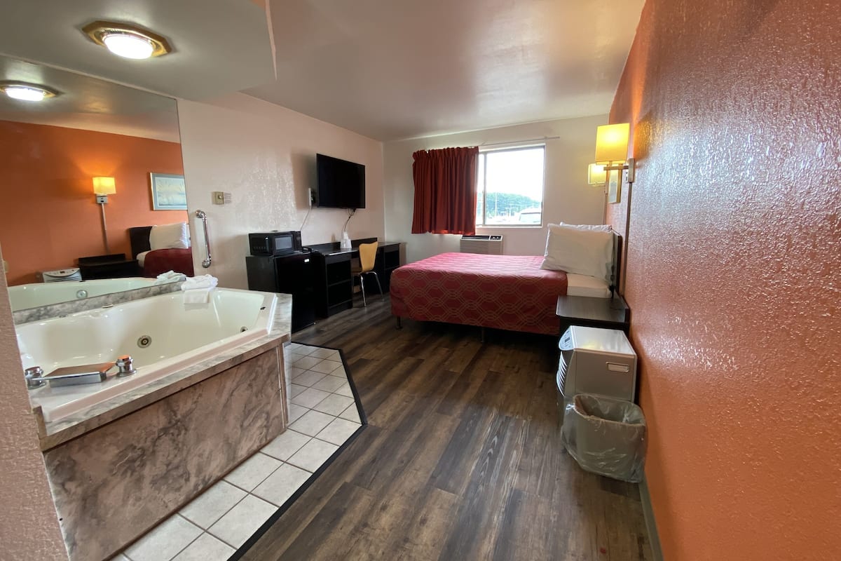 A hotel room with a large bathtub, a desk with a chair, a bed, a television, and a window with curtains.