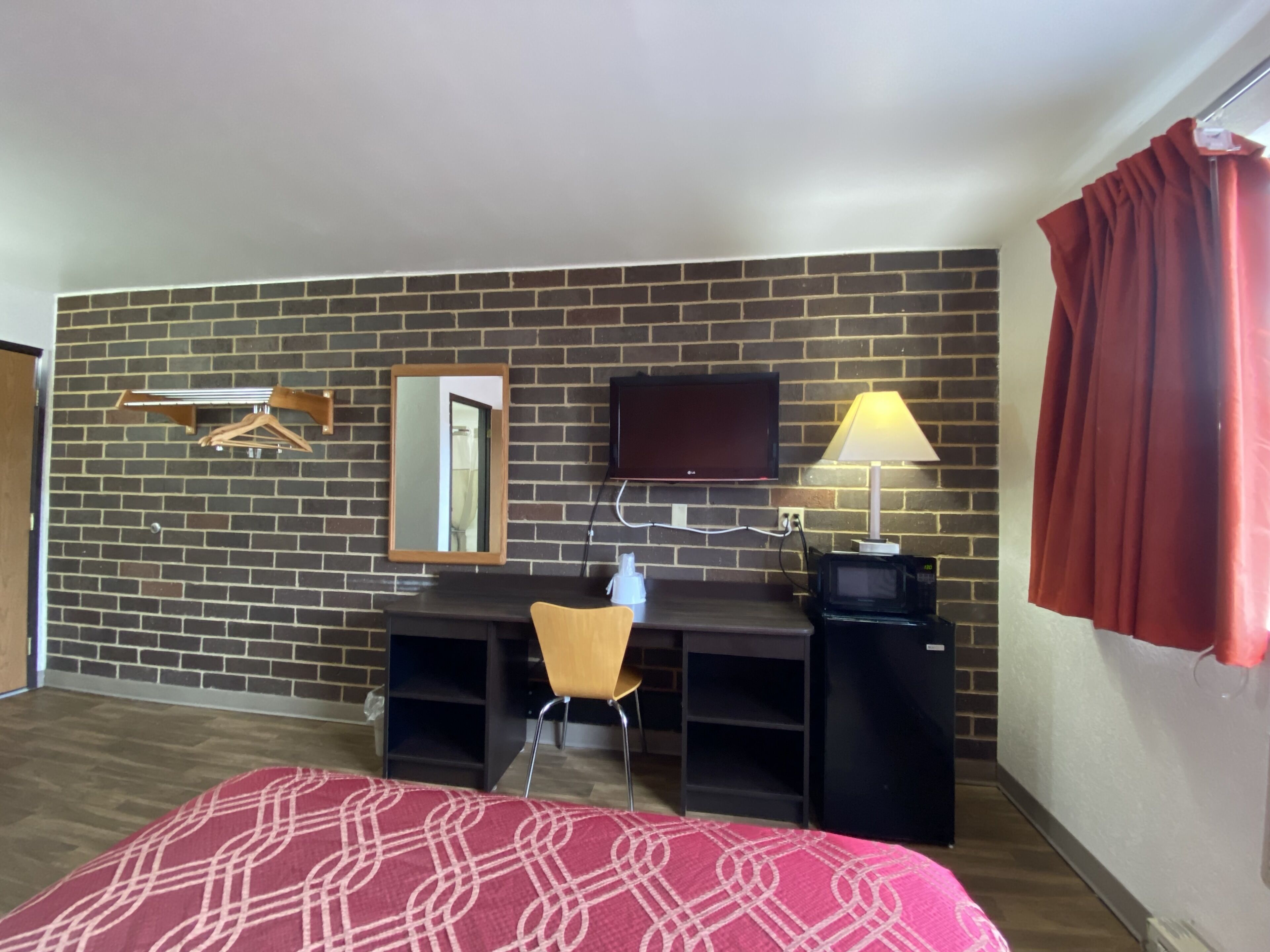 A hotel room with a brick wall, a flat-screen TV, a desk with a chair, a lamp, and a bed with a patterned bedspread.