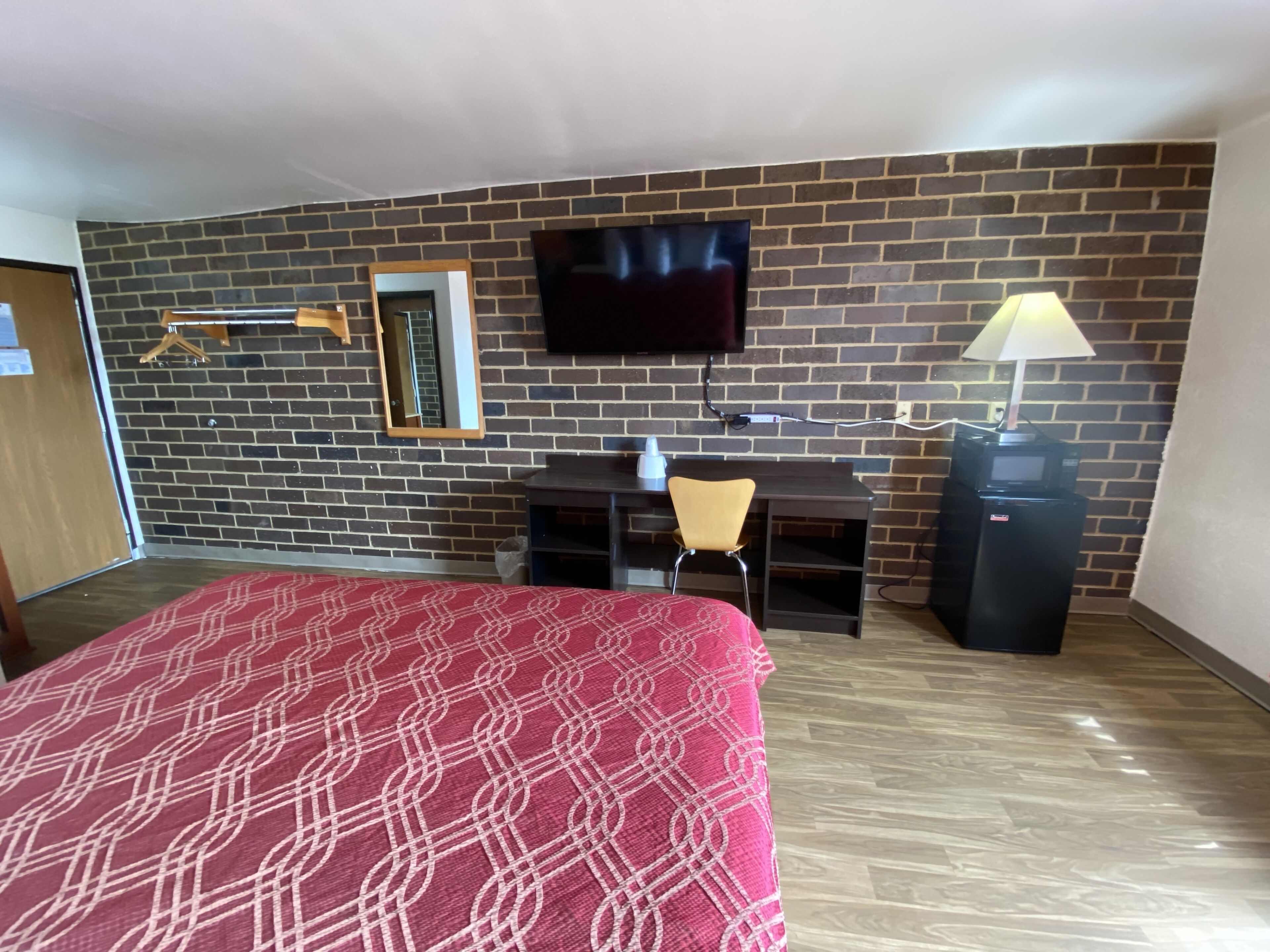 Habitación de hotel con pared de ladrillo, un televisor de pantalla plana sobre un escritorio, una silla, una cama con colcha estampada y una lámpara a la derecha.