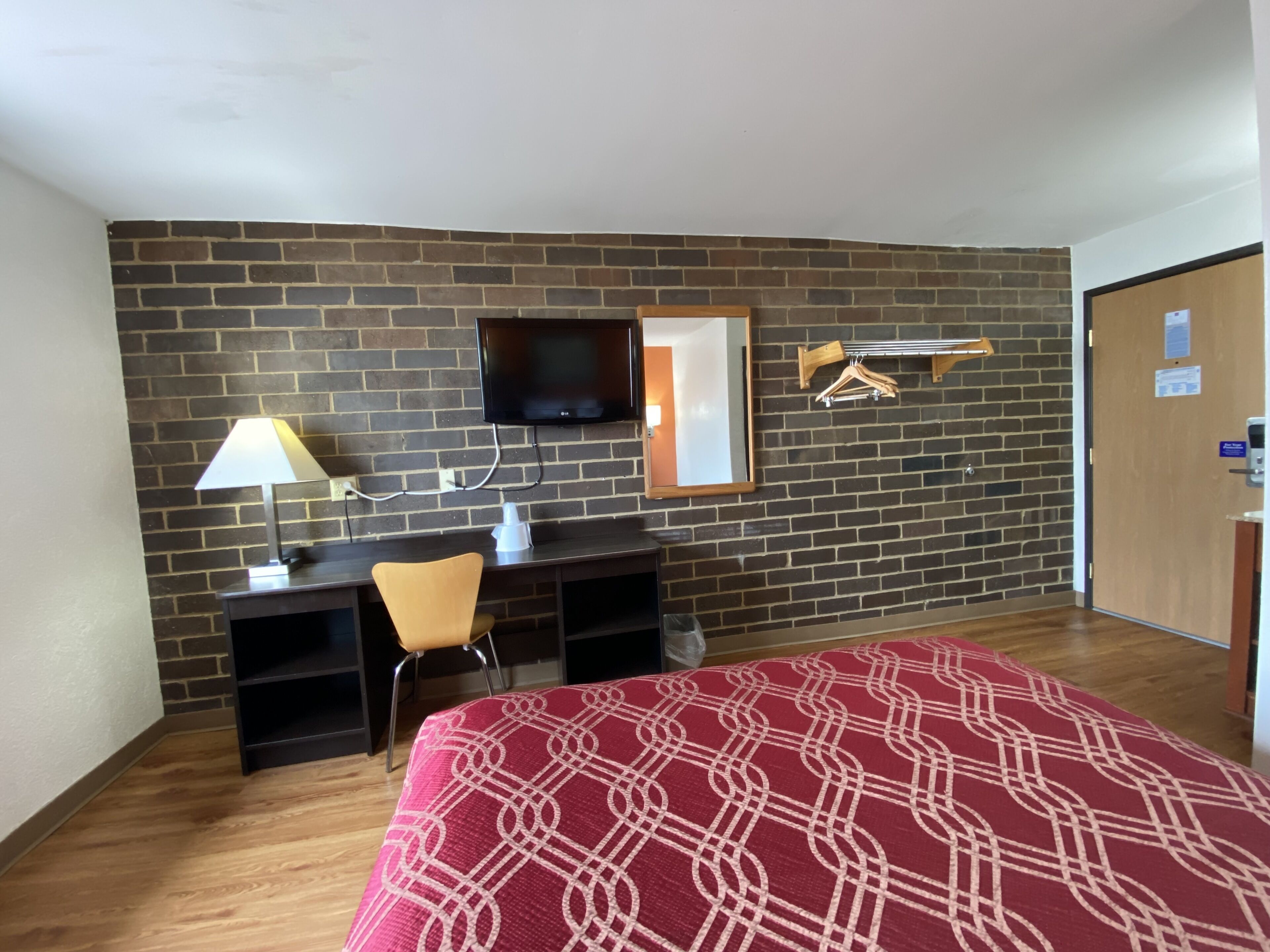 A hotel room with a brick wall, a TV mounted above a desk, a bed with a patterned bedspread, a chair, and a coat rack.