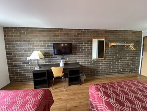 A room with a brick wall, a TV mounted above a desk, a chair, and two beds with patterned bedding.