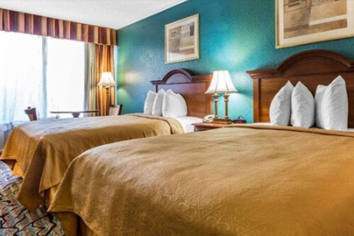 Habitación de hotel con dos camas, cabecera de madera, mesita de noche con lámpara y ventana con cortinas.
