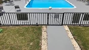 Piscina retangular com cerca de metal, um passeio pavimentado e área gramada com uma mangueira.