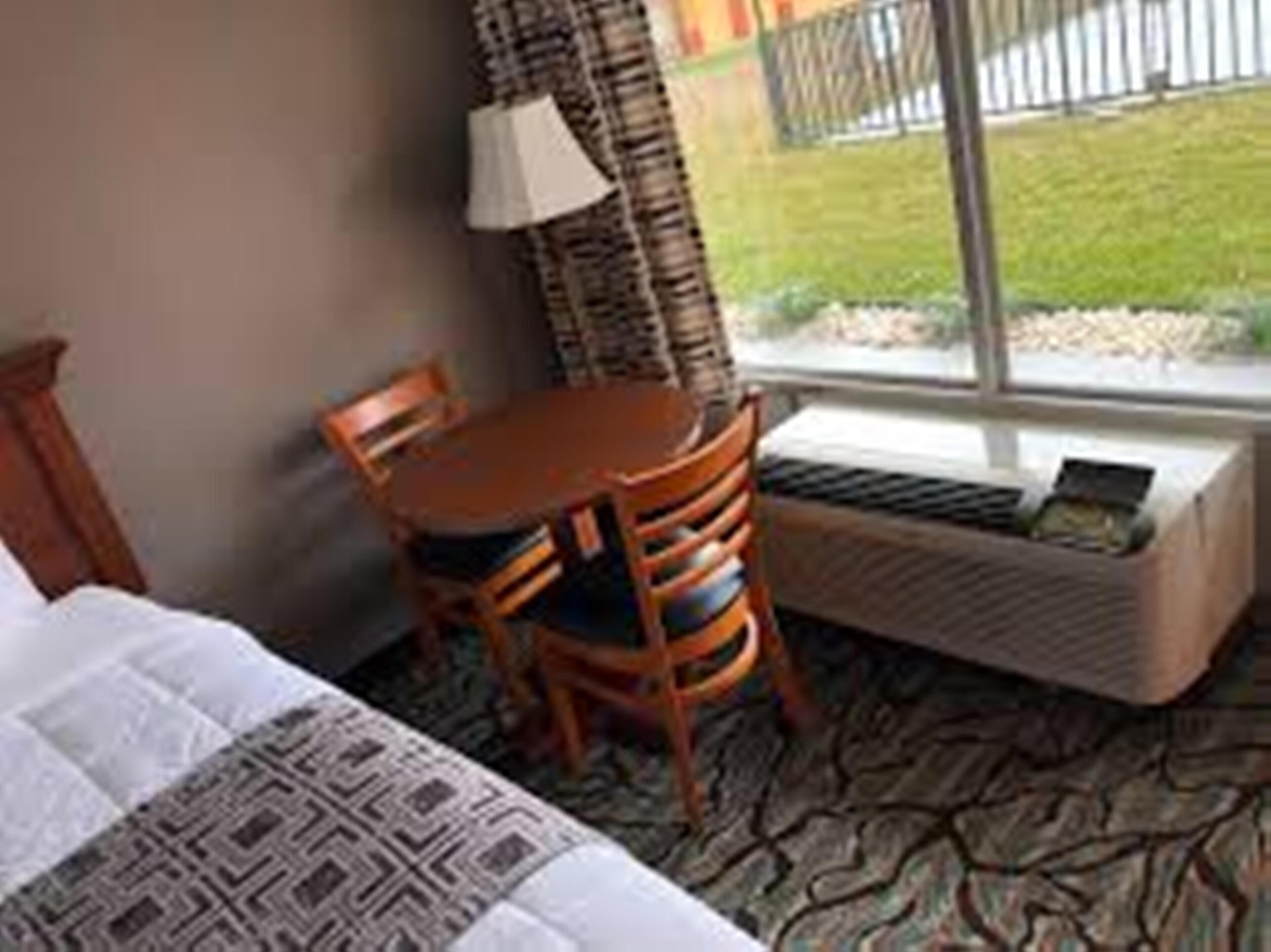 Une chambre d’hôtel avec un lit, une petite table, une chaise, une fenêtre donnant sur un espace verdoyant et un radiateur.