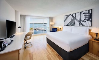 Hotelkamer met een groot bed, een bureau met een televisie, een stoel en uitzicht op een waterpartij door grote ramen.