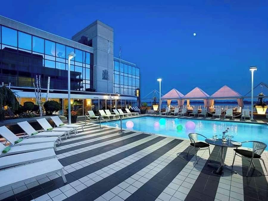 Zona de piscina moderna con sillas de descanso y sombrillas, un suelo de baldosas y un edificio grande con ventanas reflectantes.