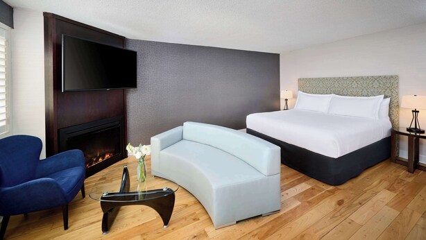A hotel room with a flat-screen TV mounted above a fireplace, a light blue sofa, a large bed with white linens, and a small table with a vase of flowers.