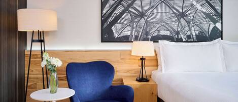 A hotel room with a bed, a blue armchair, a small round table with a vase of flowers, and two floor lamps.