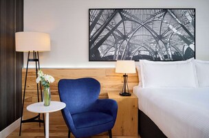 A hotel room with a bed, a blue armchair, a small round table with a vase of flowers, and two floor lamps.