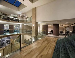 A spacious lobby with a high ceiling, a chandelier, a staircase with glass railings, and a seating area with a patterned rug.