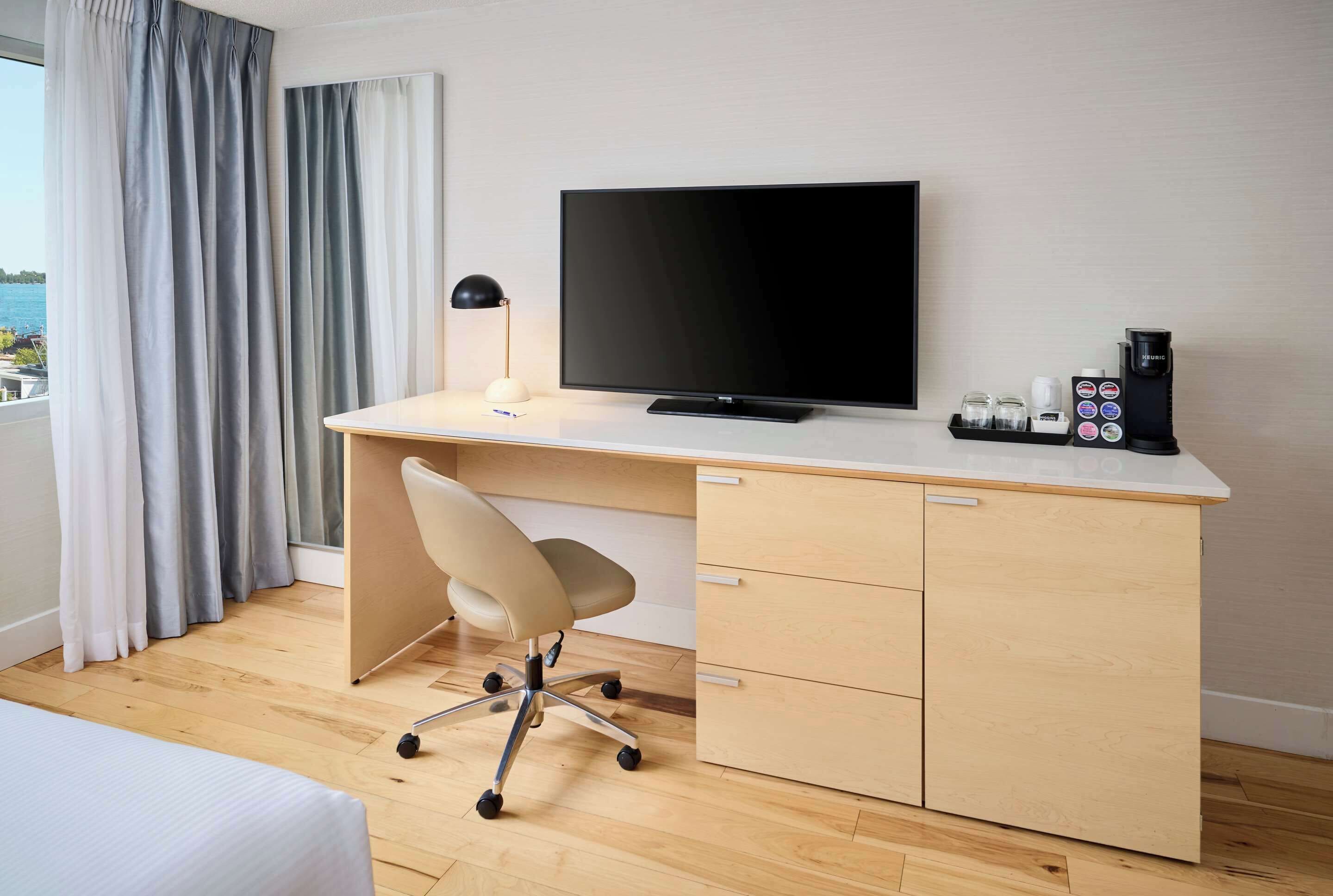 A hotel room with a wooden desk, a flat-screen TV, a desk lamp, a chair, a coffee maker, and a view of the sea through the window.