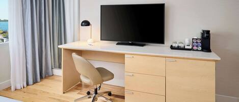 A hotel room with a wooden desk, a flat-screen TV, a desk lamp, a chair, a coffee maker, and a view of the sea through the window.